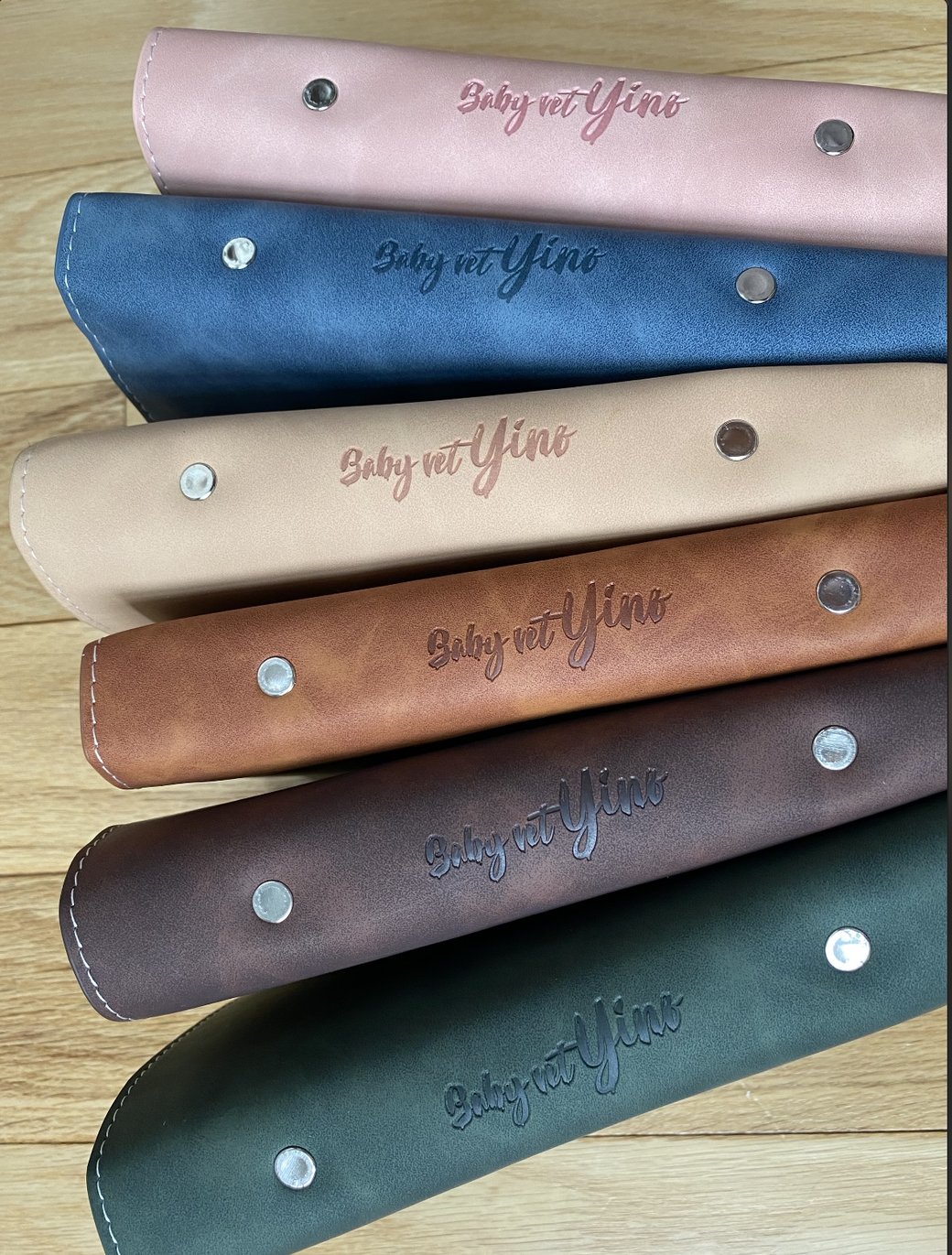 Six Baby Vet Yino handbook covers stacked — pink, blue, tan, cognac, plum, and forest green leather, each embossed with the Baby Vet Yino name